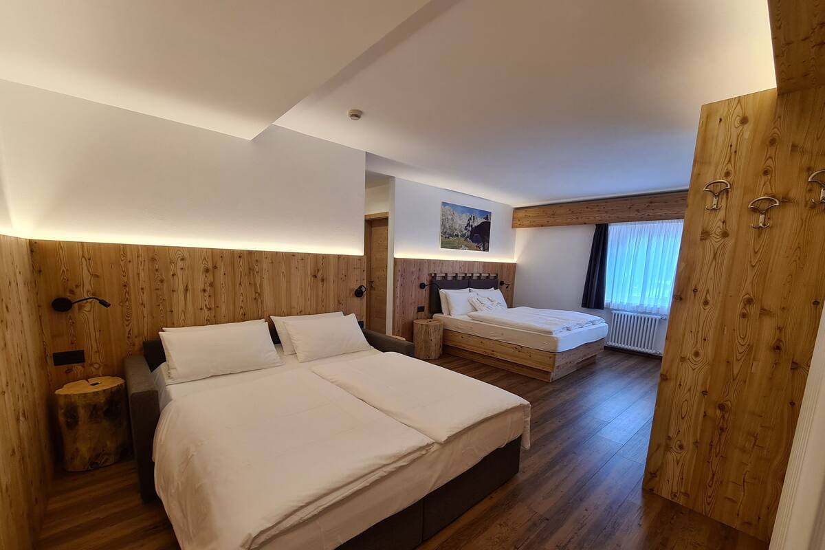 A bedroom with two beds, wooden walls, a ceiling light, a nightstand, a wooden floor, and a window with curtains.