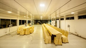 Un amplio salón de banquetes con mesas y sillas largas, ventiladores de techo y ventanas a lo largo de las paredes.