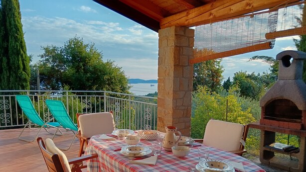 Une terrasse avec une table dressée pour le dîner, offrant une vue panoramique sur un paysage verdoyant, avec des arbres et la mer à l’horizon.