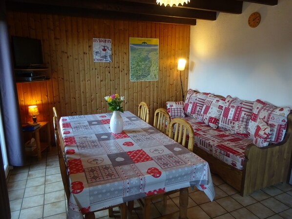 A cozy room with a wooden wall, a dining table with a quilted tablecloth, a white vase with flowers, a sofa with patterned cushions, and a television on a stand.