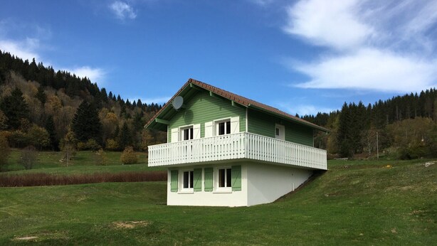 Une maison à deux étages dotée d’un balcon, entourée d’un champ verdoyant et de collines boisées.