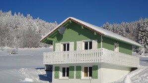 Une maison à deux étages, à l’extérieur vert et dotée d’un balcon blanc, entourée d’arbres couverts de neige.