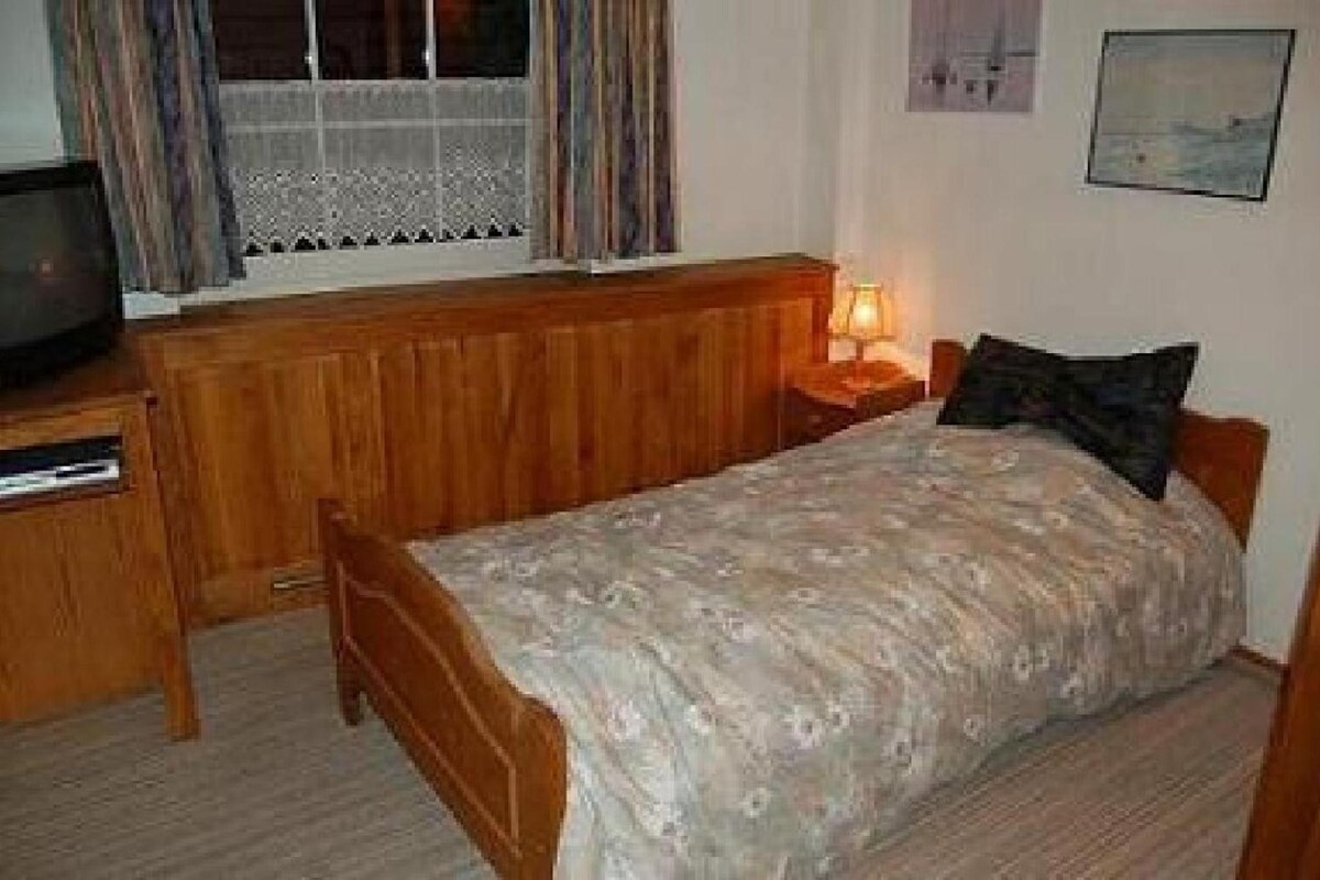 Una habitación pequeña con una cama individual, cabecera de madera, mesita de noche con lámpara y televisor sobre un aparador de madera.