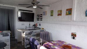 Un espacio habitable compacto con kitchenette, zona de comedor con mesa y sillas, ventilador de techo y televisor montado en la pared.