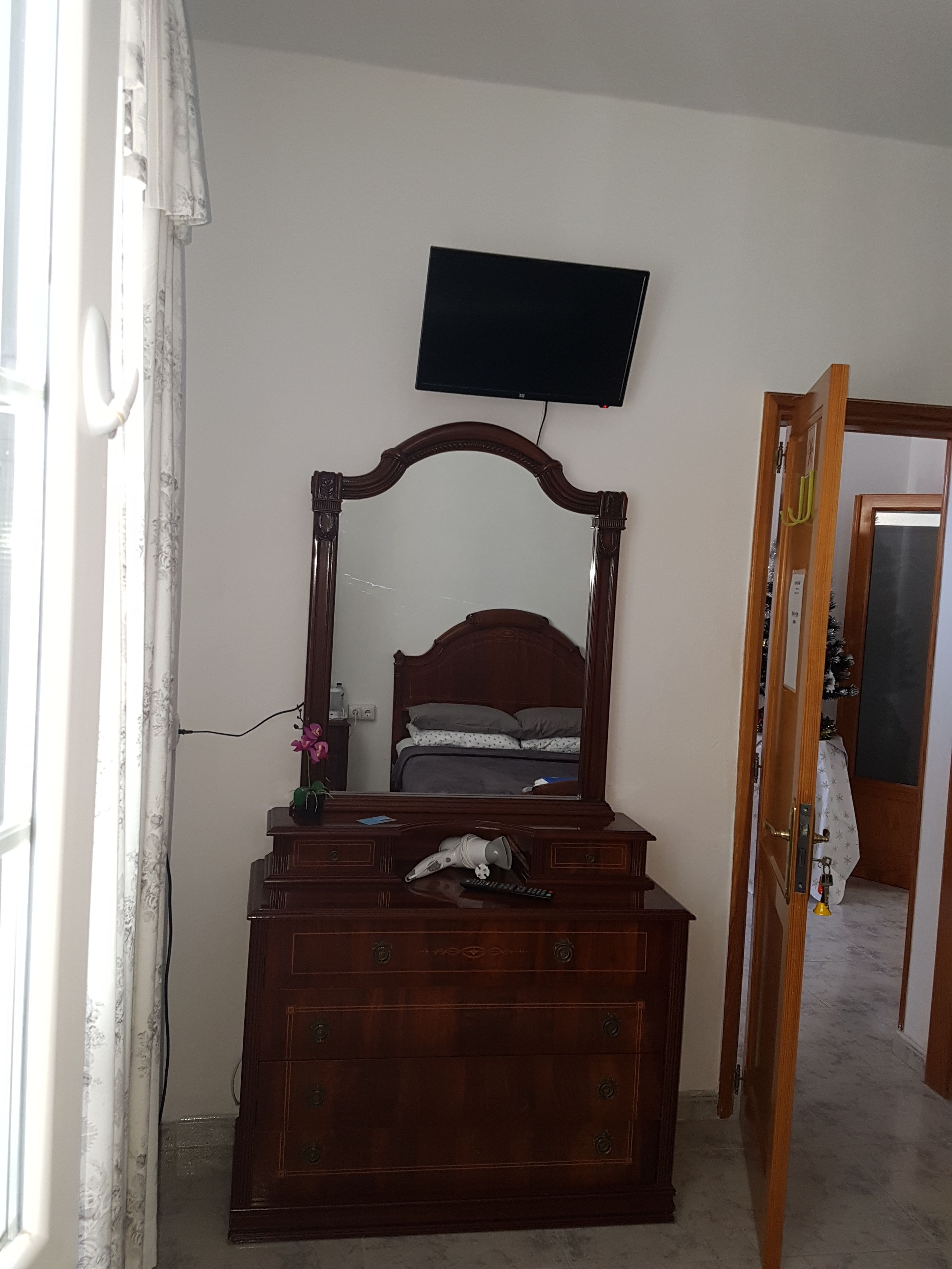 Ein Schlafzimmer mit einem Holzkleiderschrank, einem Spiegel, einem Bett mit grauer Bettwäsche, einem an der Wand montierten Fernseher und einer teilweise geöffneten Tür zu einem anderen Raum.