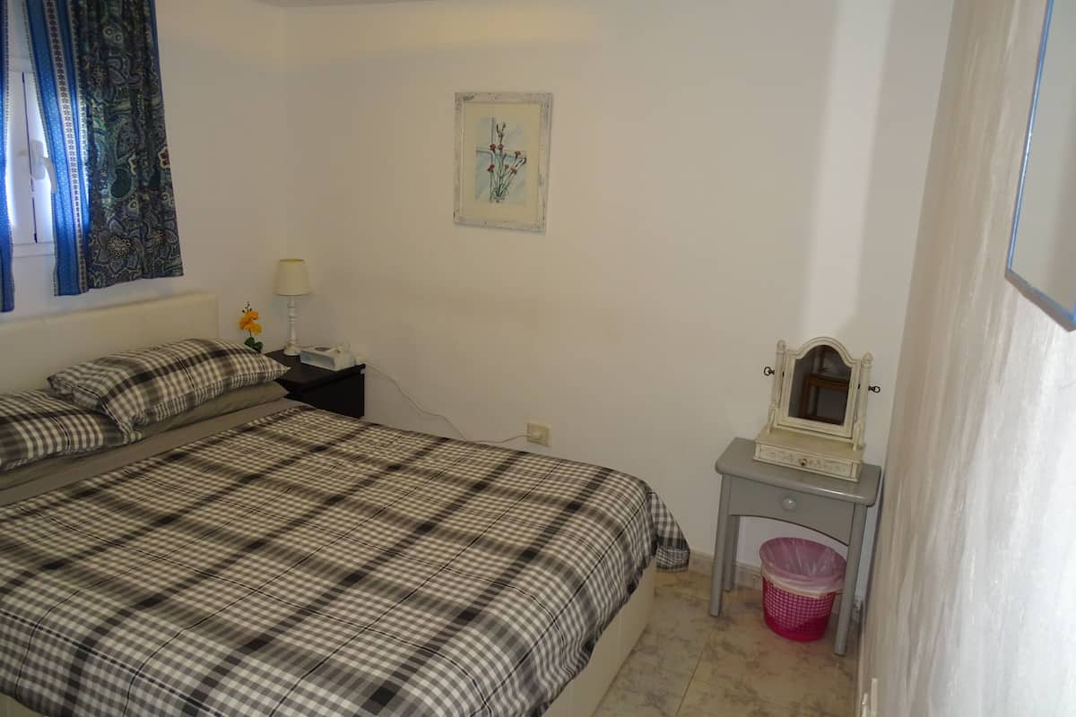 A bedroom with a large bed, a nightstand with a lamp, a small table with a mirror, and a wall-mounted picture.