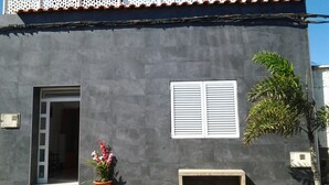 Un edificio con fachada gris, una puerta blanca, una ventana con contraventanas y una planta en maceta con flores.
