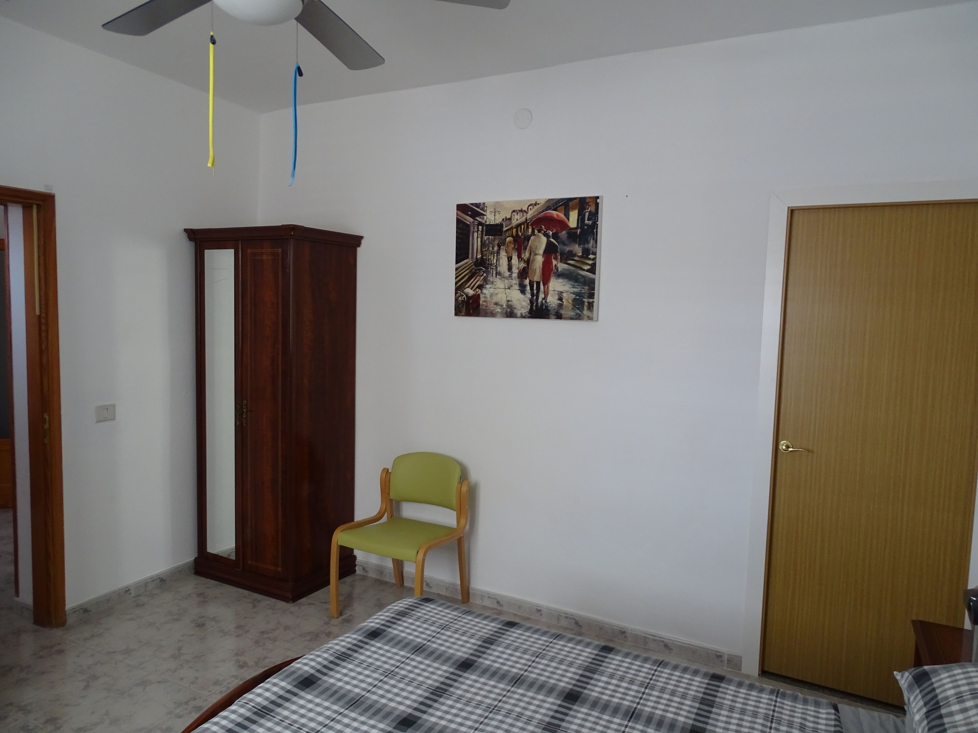 A bedroom with a bed, a wooden wardrobe, a green chair, a door, and a wall-mounted painting.