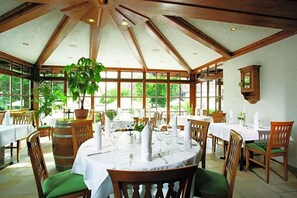 A dining area with tables set for a meal, wooden beams on the ceiling, and large windows with greenery outside.