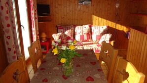 Een gezellige kamer met houten wanden, een bank met gestreepte kussens, een eettafel met een bloemstuk en een televisie op een standaard.