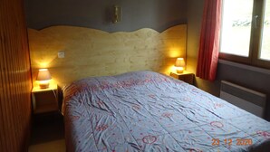 Een slaapkamer met een houten hoofdeinde, twee bedlampen, een bed met een beddengoed met patroon, en een raam met een rood gordijn.