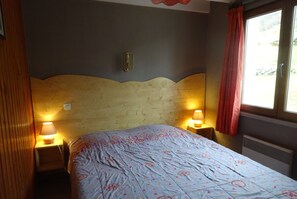 Een slaapkamer met een houten hoofdeinde, twee bedlampen, een bed met een beddengoed met patroon, en een raam met een rood gordijn.