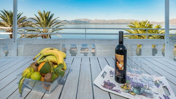 A table with a bottle of wine, a fruit bowl, and a placemat with glasses on a balcony overlooking a body of water and mountains. - Beautiful apartment for 4 guests with A/C, WIFI, TV and patio (Alcúdia)