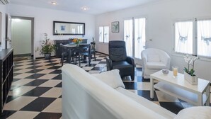 A living room with a checkered floor, a sofa, a dining table with chairs, a black armchair, a white armchair, and a small table with a candle and a plant. - Beautiful apartment for 4 guests with A/C, WIFI, TV and patio (Alcúdia)