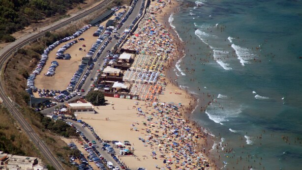Vista aérea de una playa con filas de coches aparcados, sombrillas y gente disfrutando del paseo marítimo.
