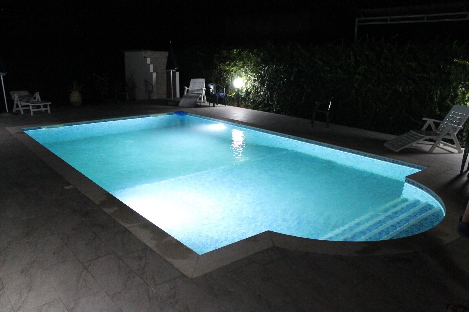 A rectangular swimming pool with a tiled edge, surrounded by lounge chairs and a small building in the background.
