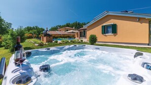 A hot tub with a bottle of wine, surrounded by a lush garden and a house with a swimming pool in the background.