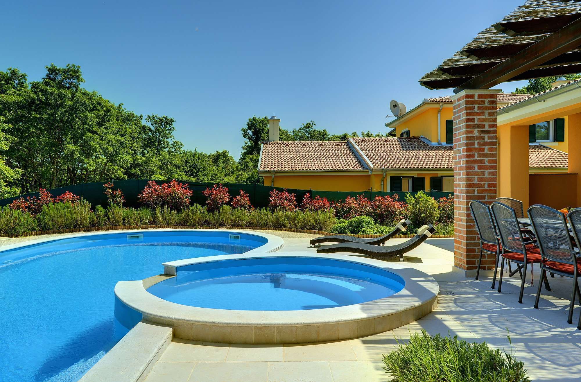 Une piscine à bord incurvé, un espace détente avec des fauteuils et, au fond, un bâtiment avec un toit recouvert de tuiles.