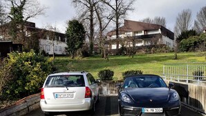 A parking area with two cars, a white hatchback and a black sports car, parked on a paved surface with a grassy area and buildings in the background. - Fantastic, exclusive apartment with fantastic lake and alpine views (Immenstaad am Bodensee)