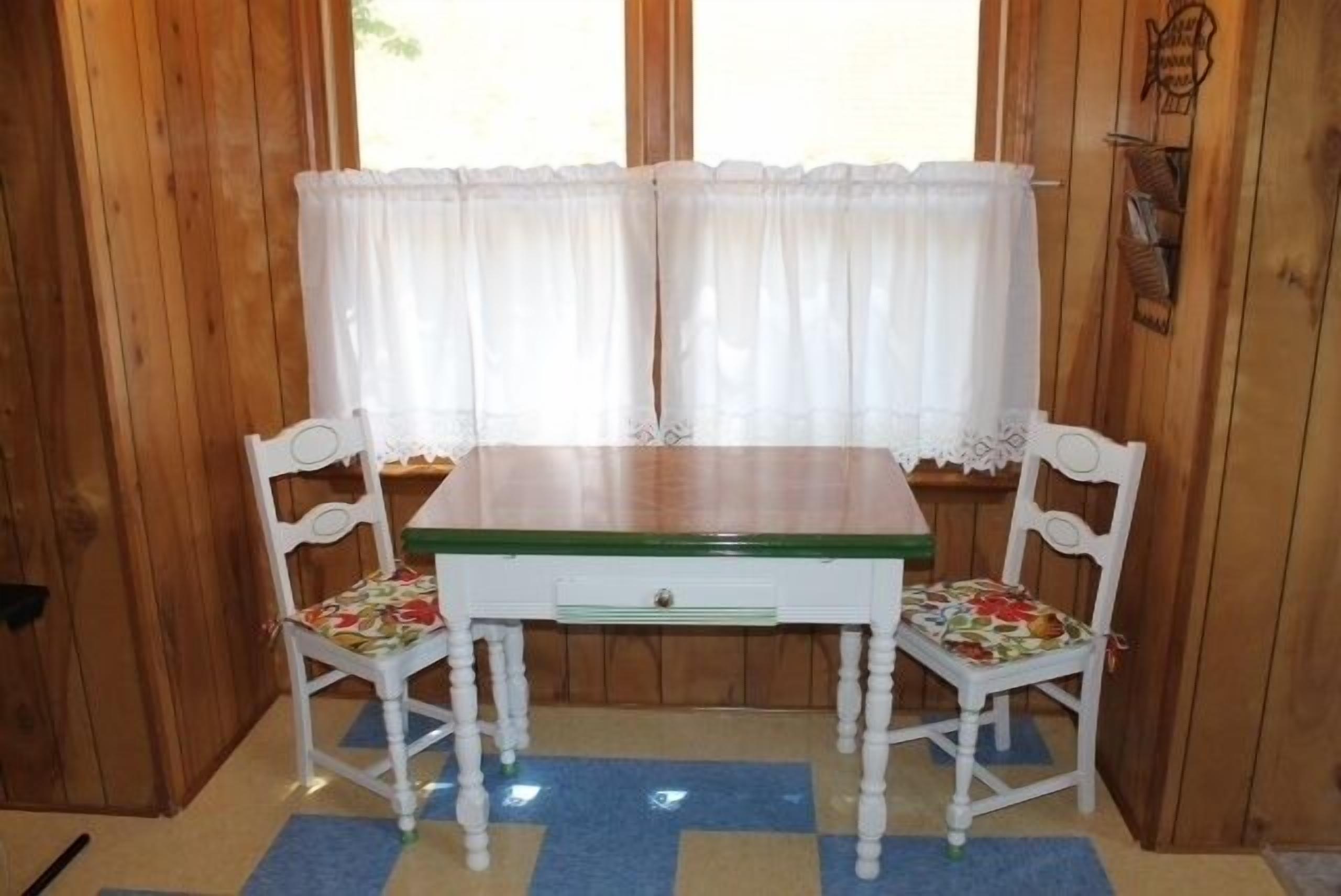 A wooden room with a white table and two chairs, a window with sheer curtains, and a wooden wall with a fish decoration.