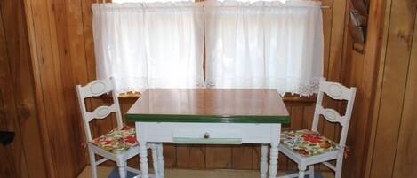 A wooden room with a white table and two chairs, a window with sheer curtains, and a wooden wall with a fish decoration.