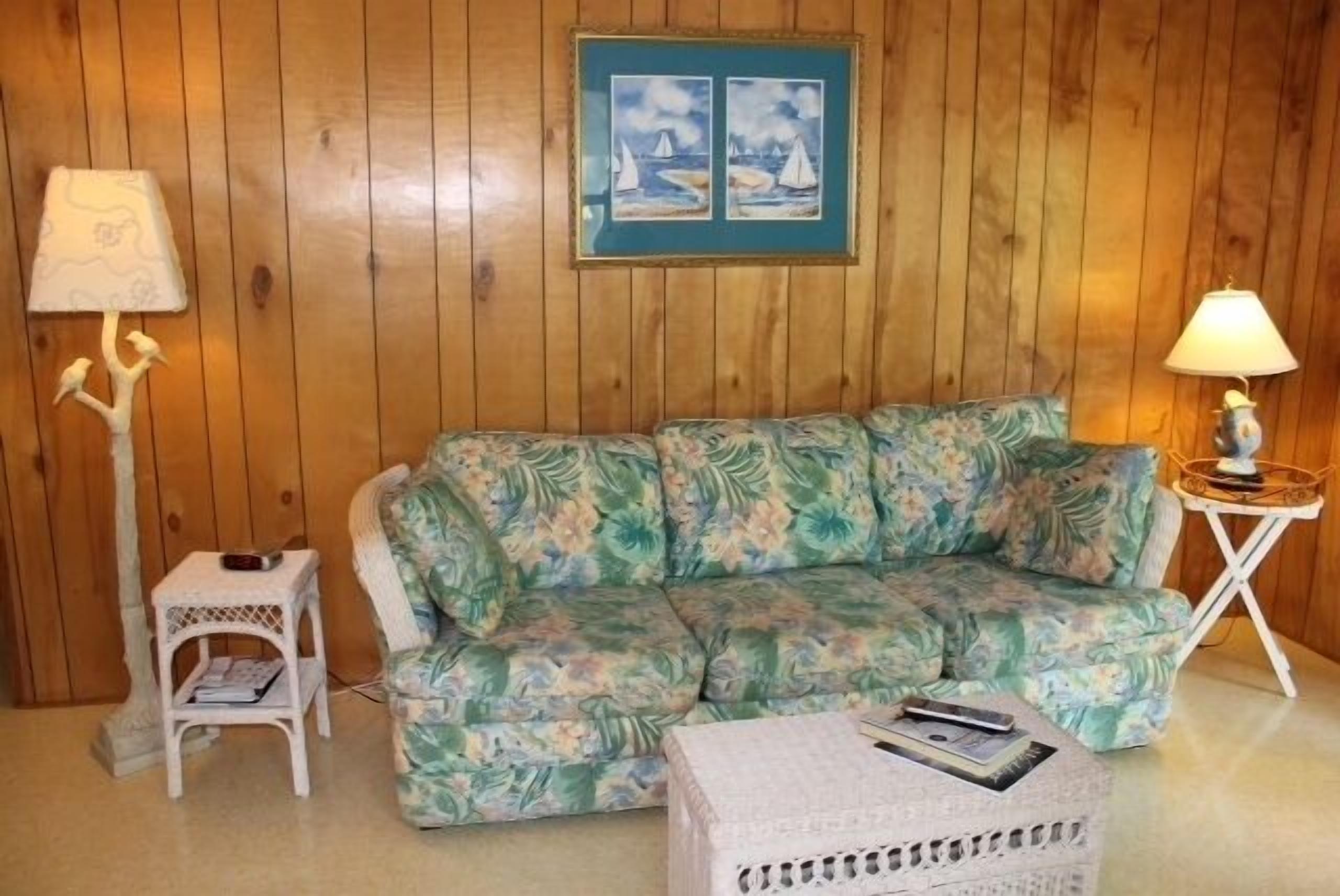 Ein Zimmer mit Holzwänden, einem sofa mit floralem Muster, zwei Nachttischen mit Lampen, einem gerahmten Bild an der Wand und einem kleinen weißen Beistelltisch mit einer Zeitschrift.