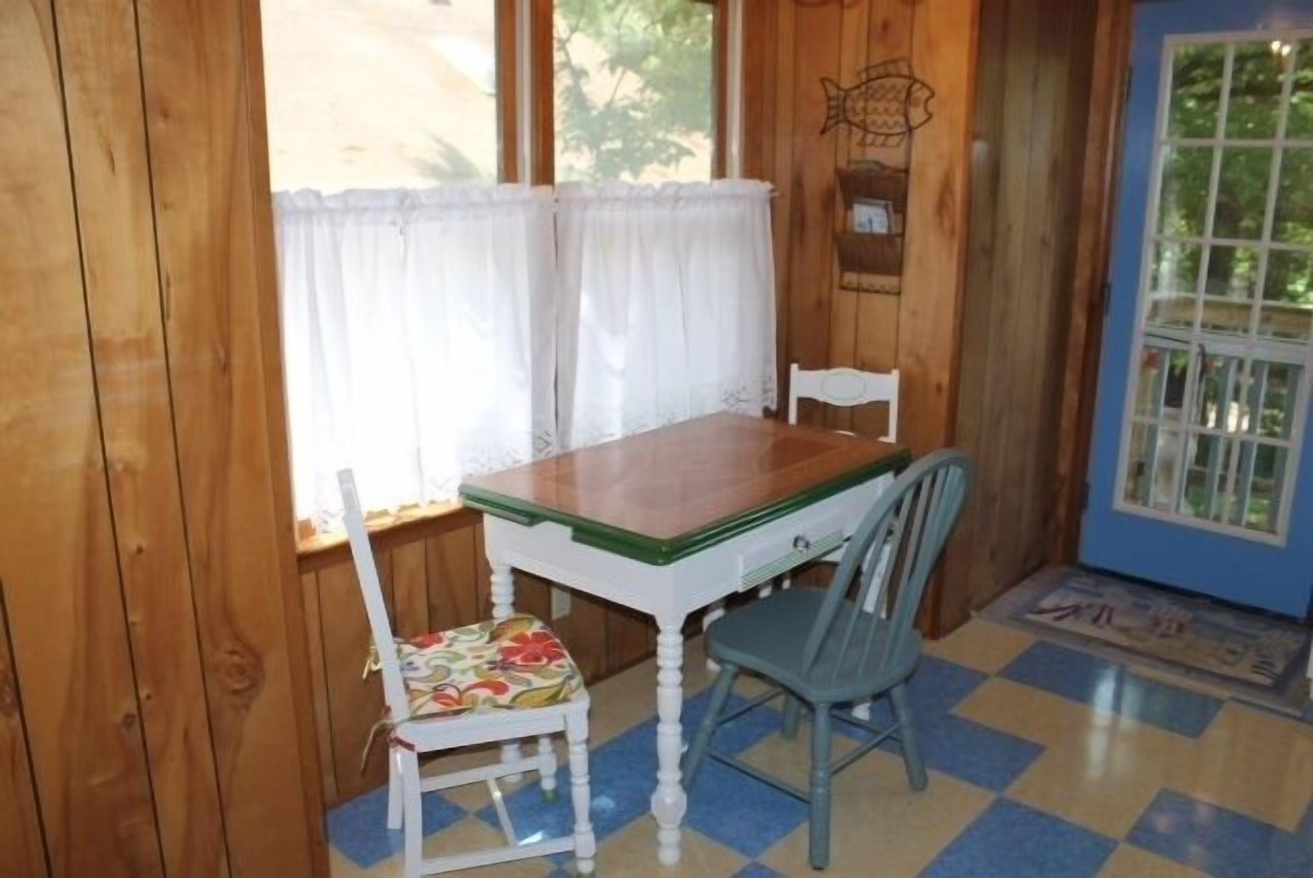 Een houten interieur met een tafel en twee stoelen, een raam met gordijnen en een blauw-geel geruit vloertje.