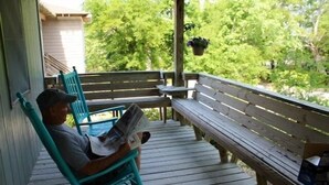 A wooden deck with a bench and a chair, a person reading a newspaper, and a hanging plant. - Treehouse OBX. Cozy cottage in family/pet-friendly neighborhood. Close to sound. (Kill Devil Hills)