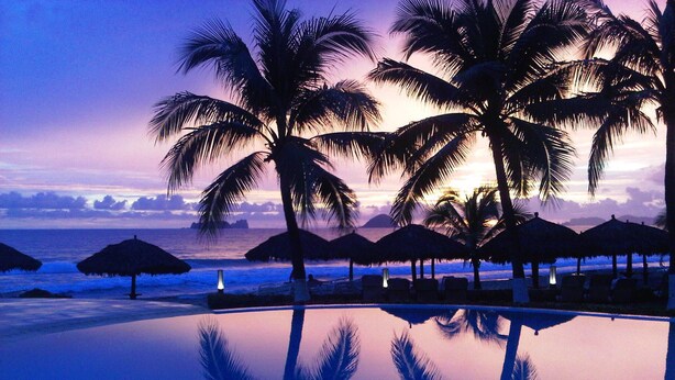 Vista a la piscina con palmeras y sombrillas de paja, reflejando la puesta de sol sobre el océano.