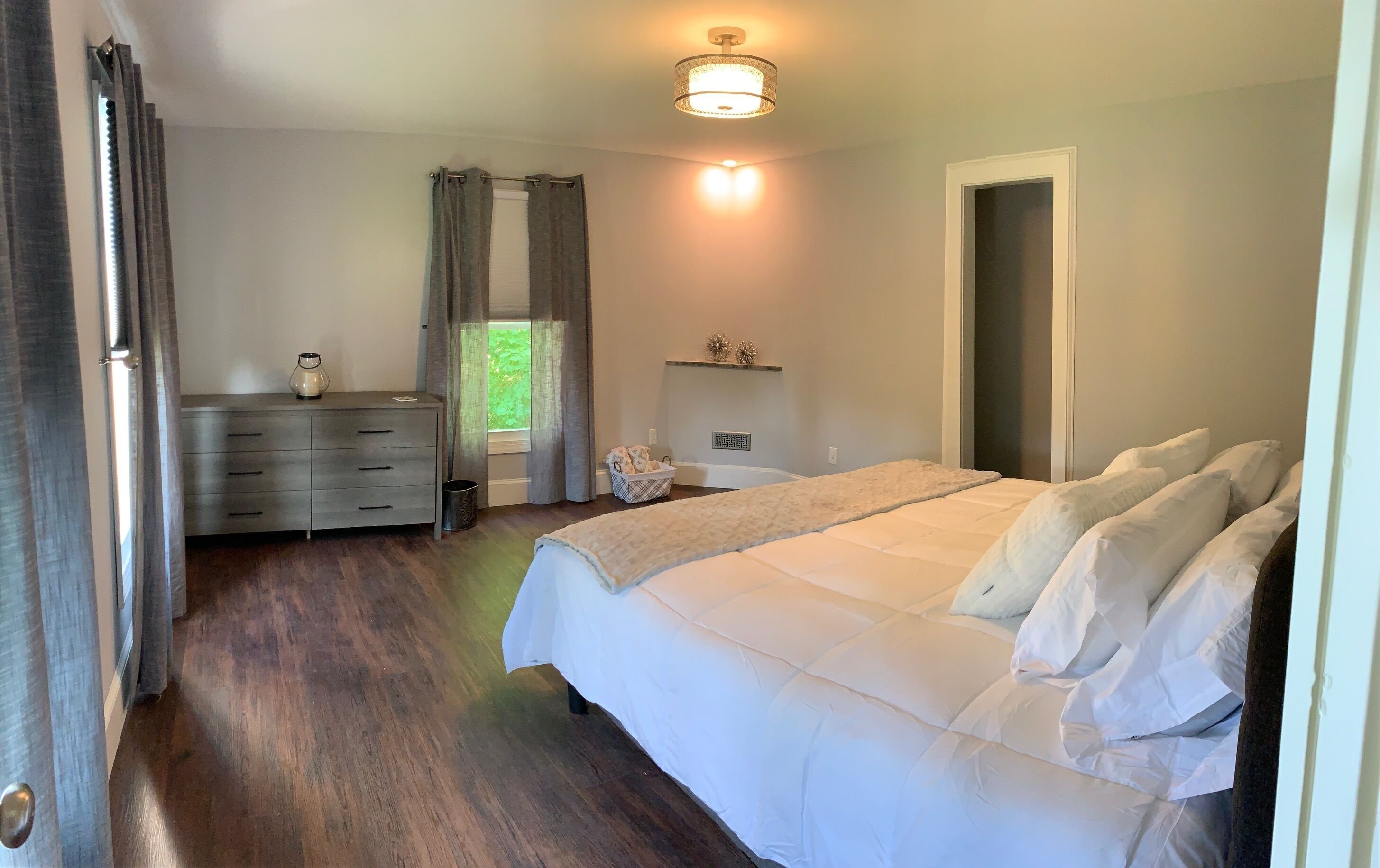 Une chambre à coucher avec un grand lit, une commode, une fenêtre avec des rideaux, un luminaire au plafond et une porte.