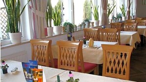 A dining area with tables set for two, wooden chairs, potted plants, and a window with curtains.