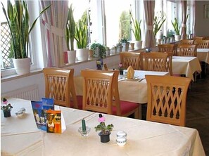 A dining area with tables set for two, wooden chairs, potted plants, and a window with curtains.