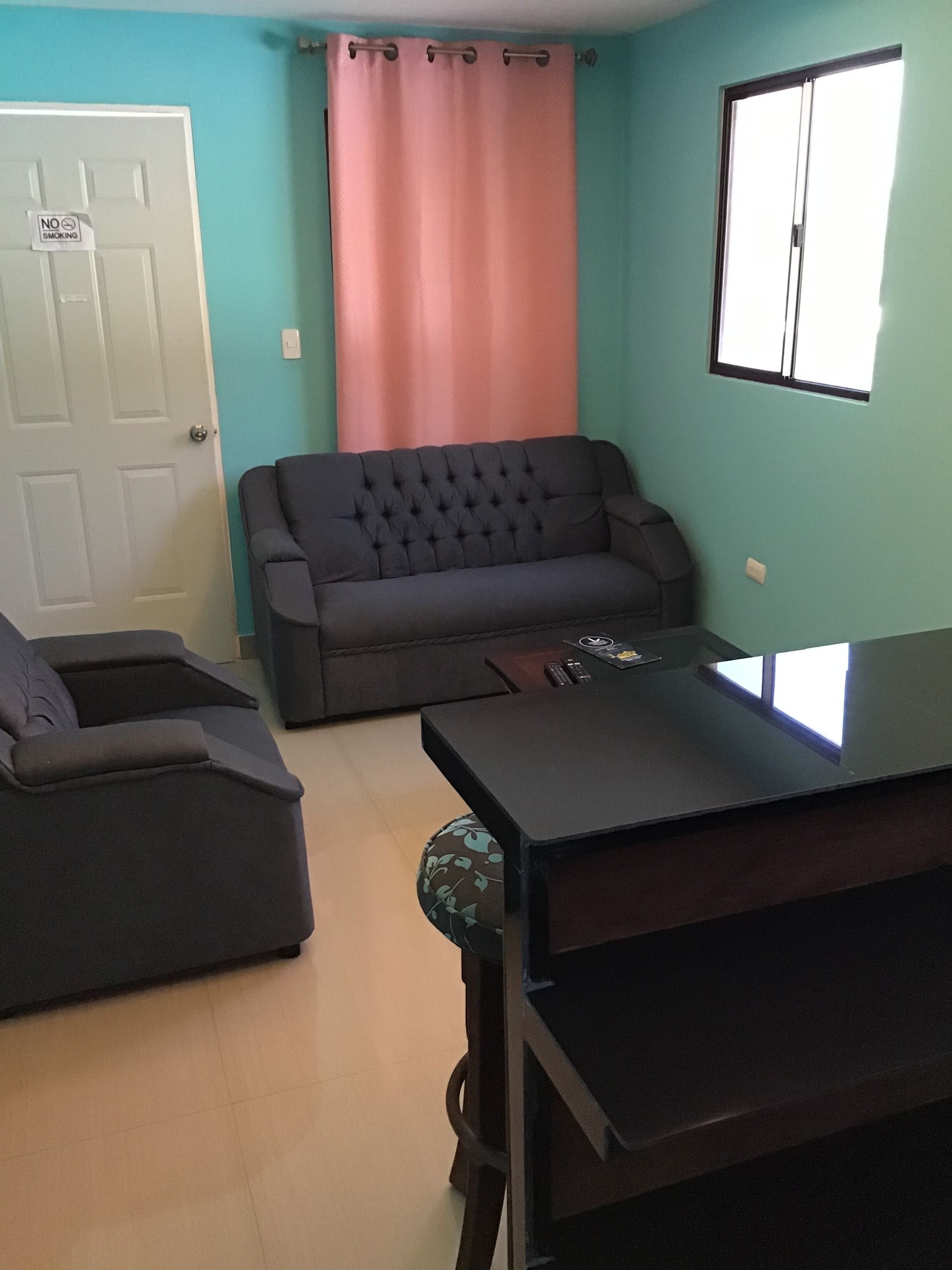 A living room with a dark grey sofa, a matching armchair, and a coffee table. A door with a 'No Smoking' sign is visible, and a window with blinds is on the right.