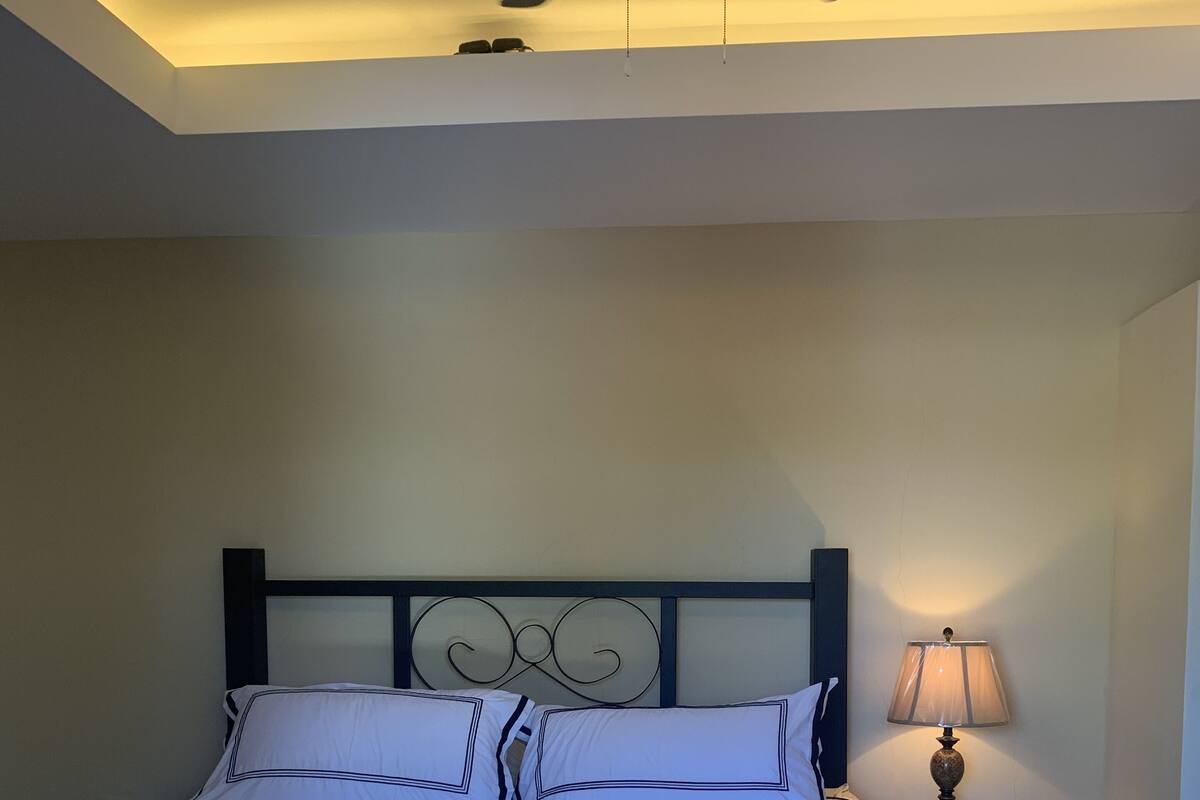 A neatly made bed with white linens and a dark headboard, a bedside lamp, and a ceiling fan.