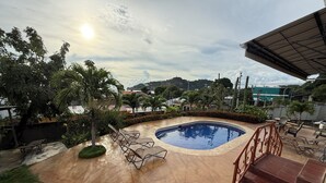 Ein Pool mit Liegestühlen, umgeben von Grünpflanzen und Blick auf ferne Hügel.