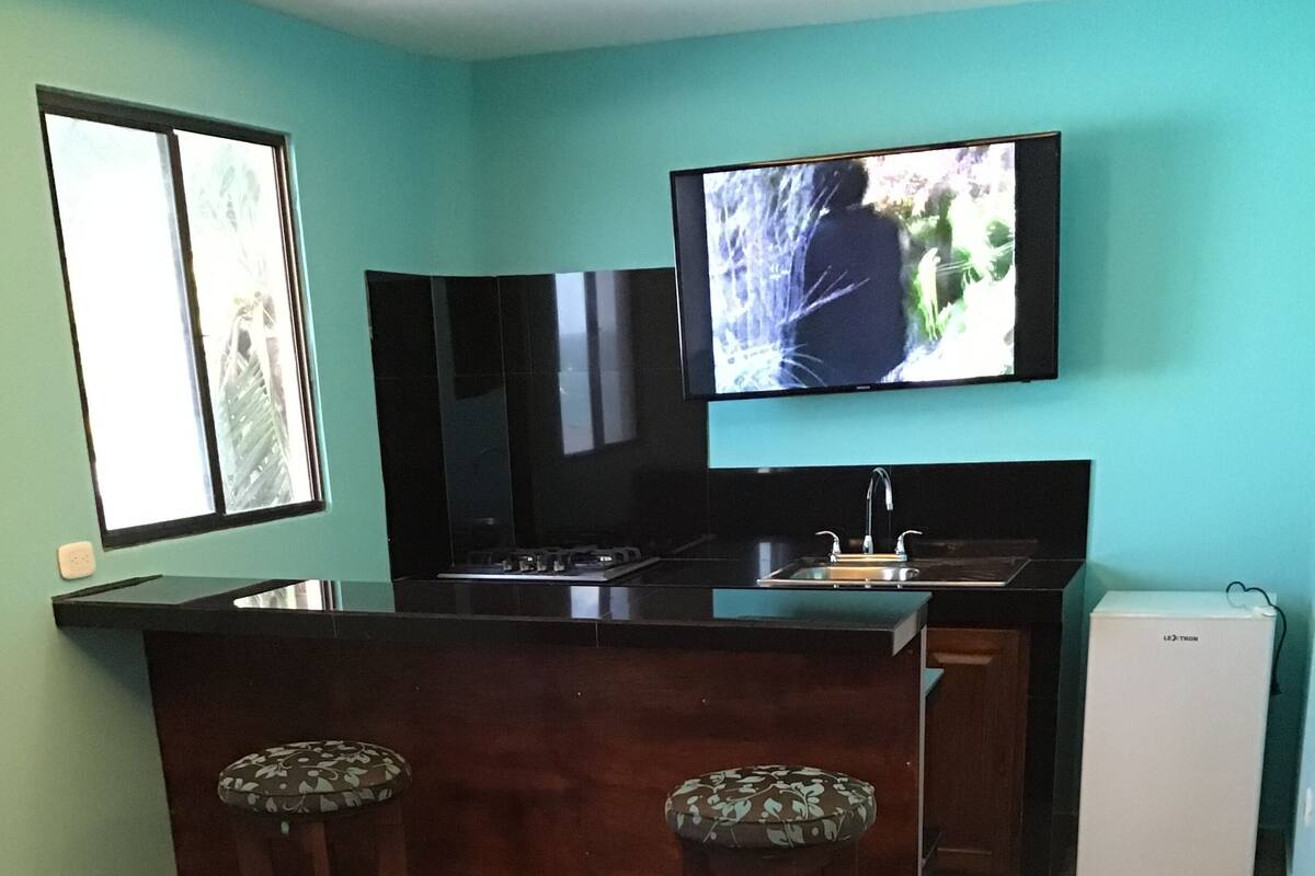 A modern kitchen with a central island, two bar stools, a refrigerator, and a flat-screen TV mounted on the wall.