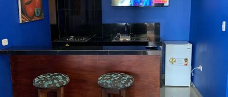 Une cuisine avec des murs bleus, un plan de travail noir, deux tabourets, une télévision et un réfrigérateur.