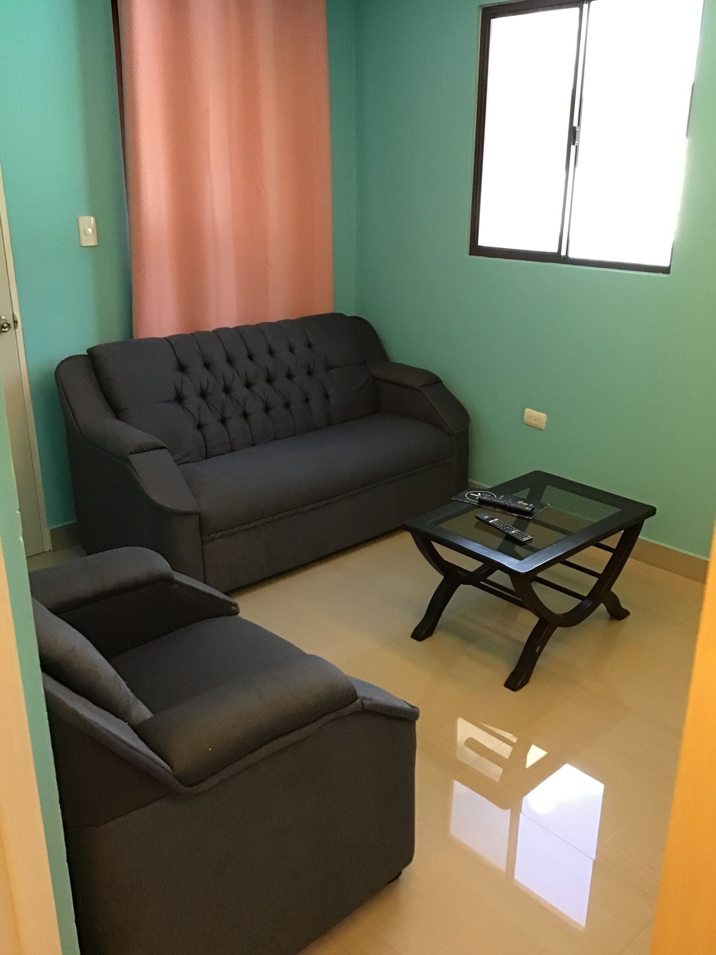 Un salon avec un canapé de couleur sombre, une table basse en verre et une fenêtre équipée de stores.