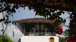 A white building with a tiled roof and a small balcony, surrounded by lush greenery and flowering plants. - Hotel Hacienda Supracafe (Cajibío)