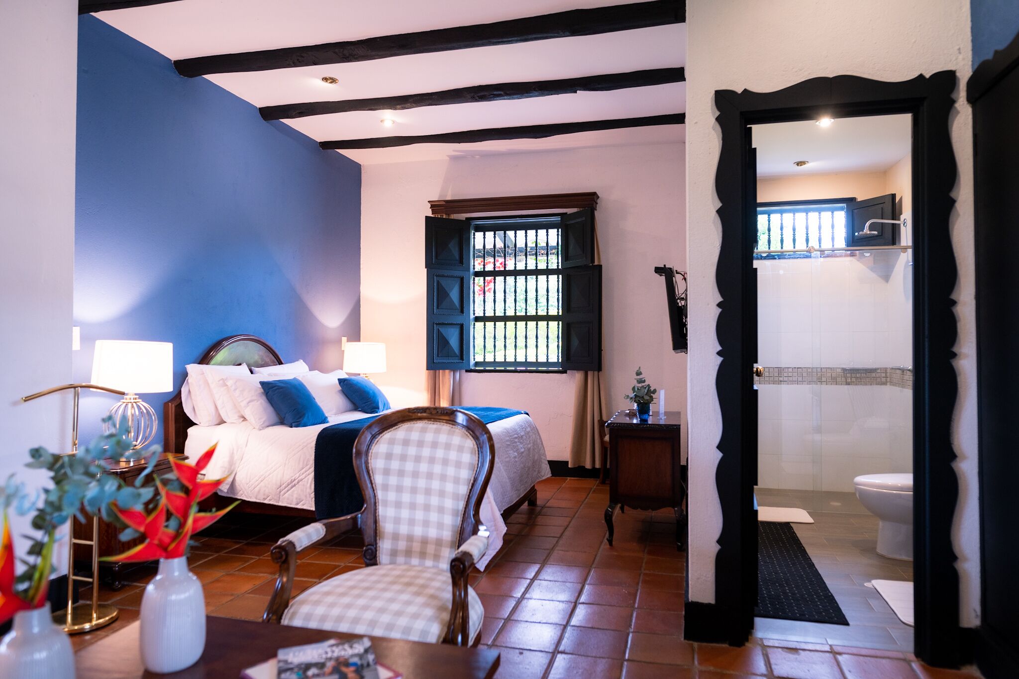 A hotel room with a bed, a chair, a nightstand, a window with bars, and a bathroom visible through an open door.
