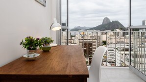 A modern room with a wooden table, two potted plants, a white chair, and a large window offering a cityscape view.