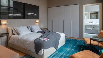 Quarto moderno com cama, mesas de cabeceira, um tapete azul e um espaço de estar com poltrona e mesa.