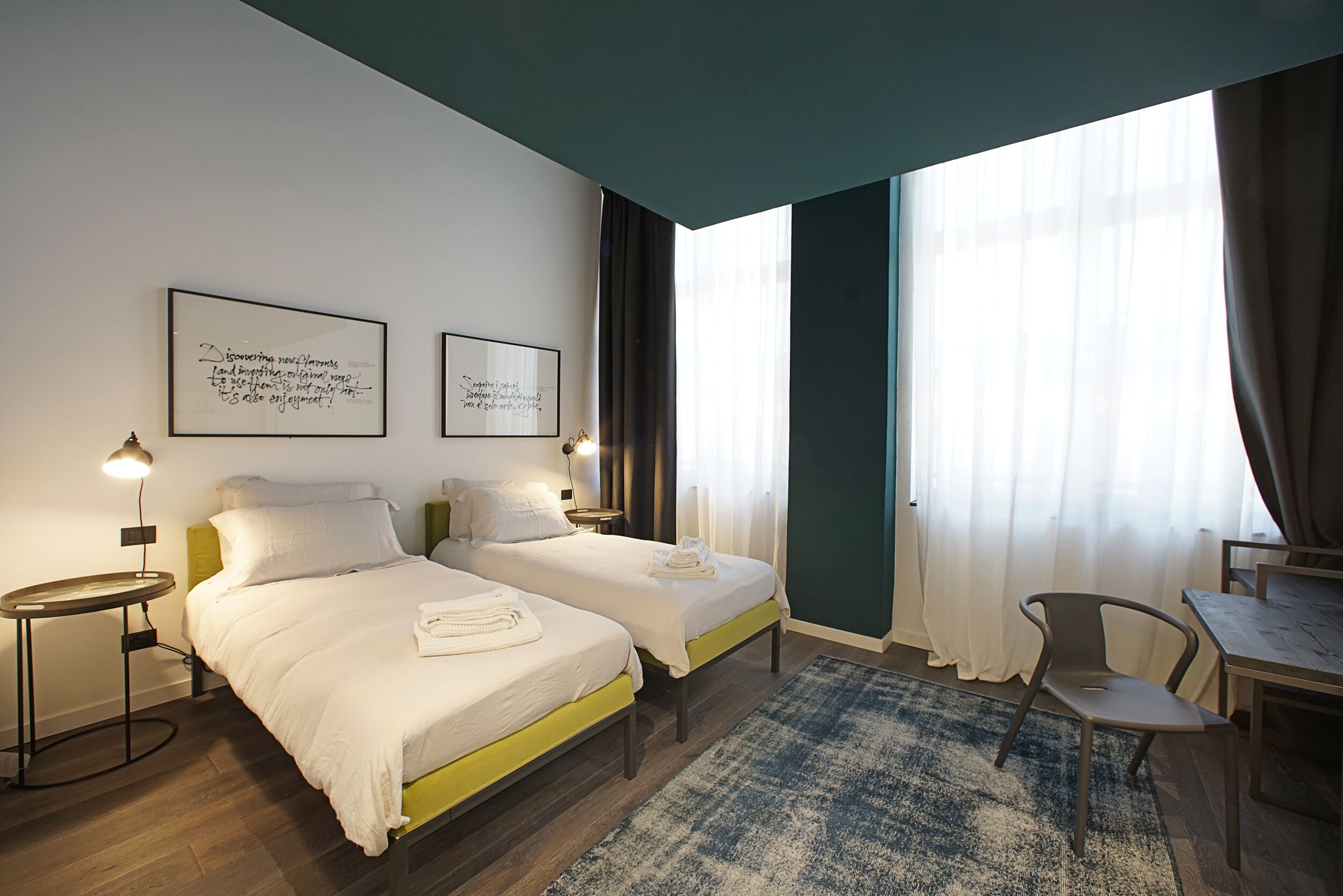 Een hotelkamer met twee bedden, een bureau en een stoel. Er hangen prestaties aan de muur en een raam met gordijnen.