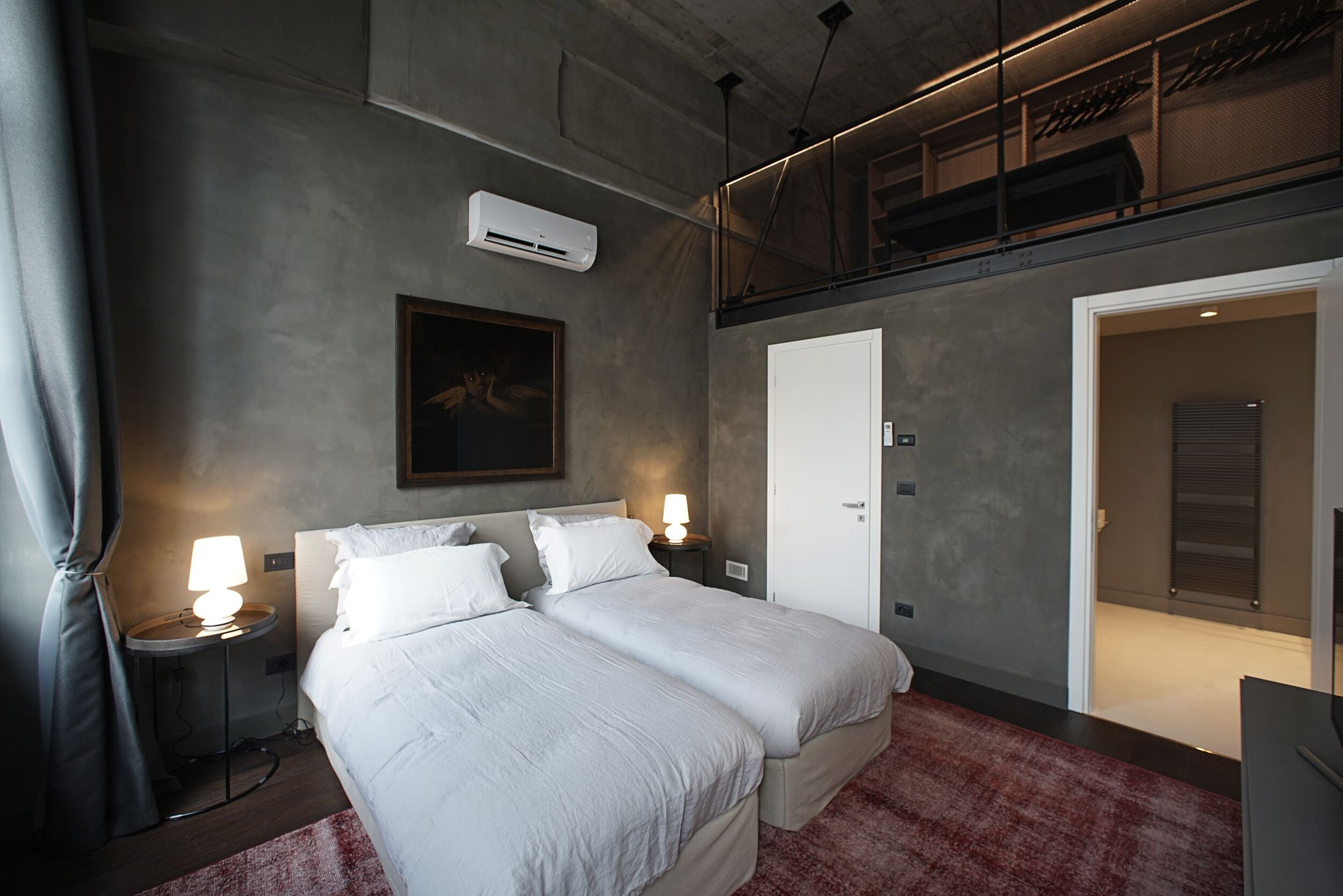 A bedroom with a large bed, two bedside tables with lamps, an air conditioning unit, a framed picture on the wall, and a door leading to another room.