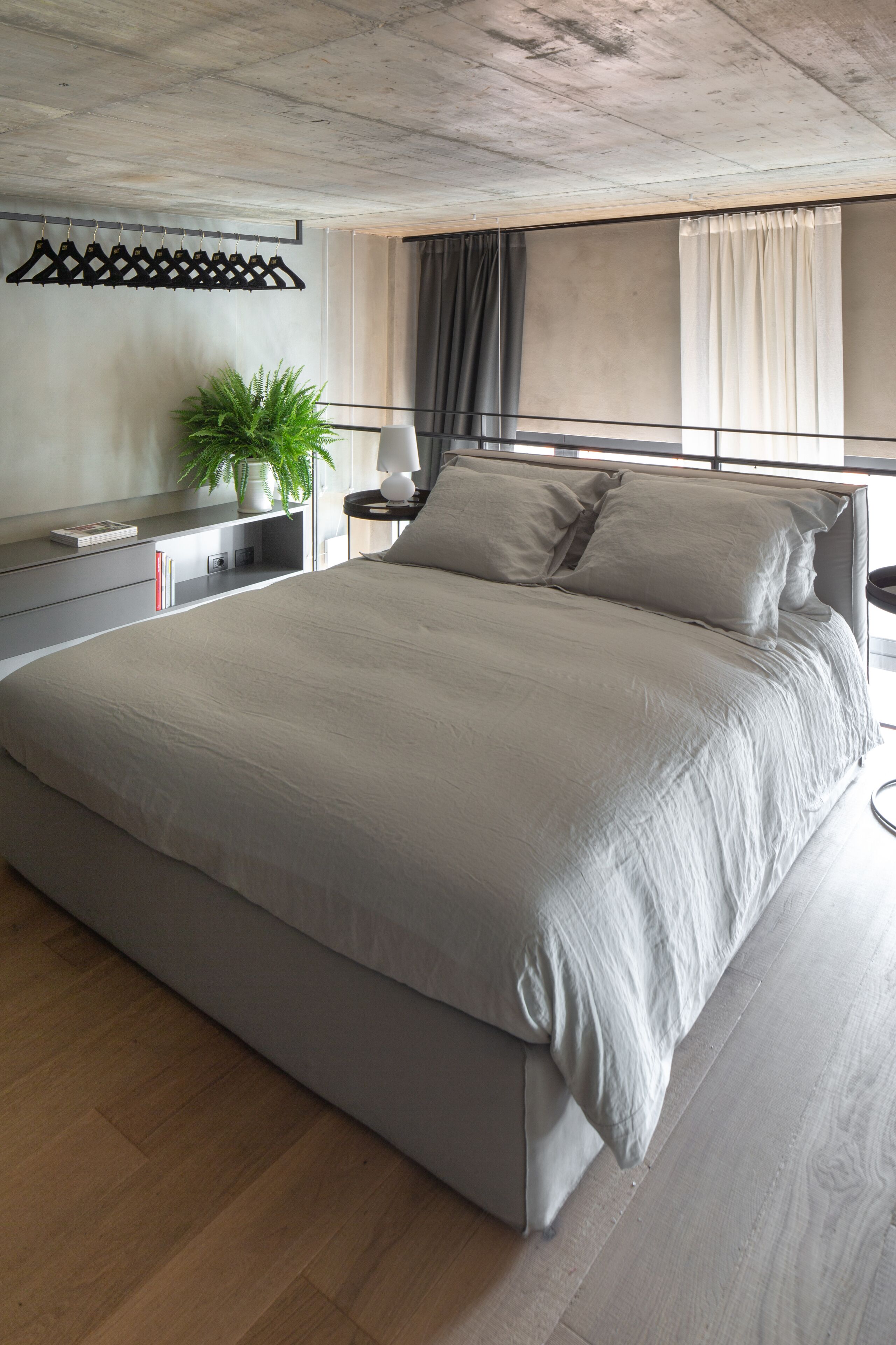 Un dormitorio moderno con una cama grande, mesitas de noche, una planta en maceta y vistas de la ciudad a través de amplios ventanales.