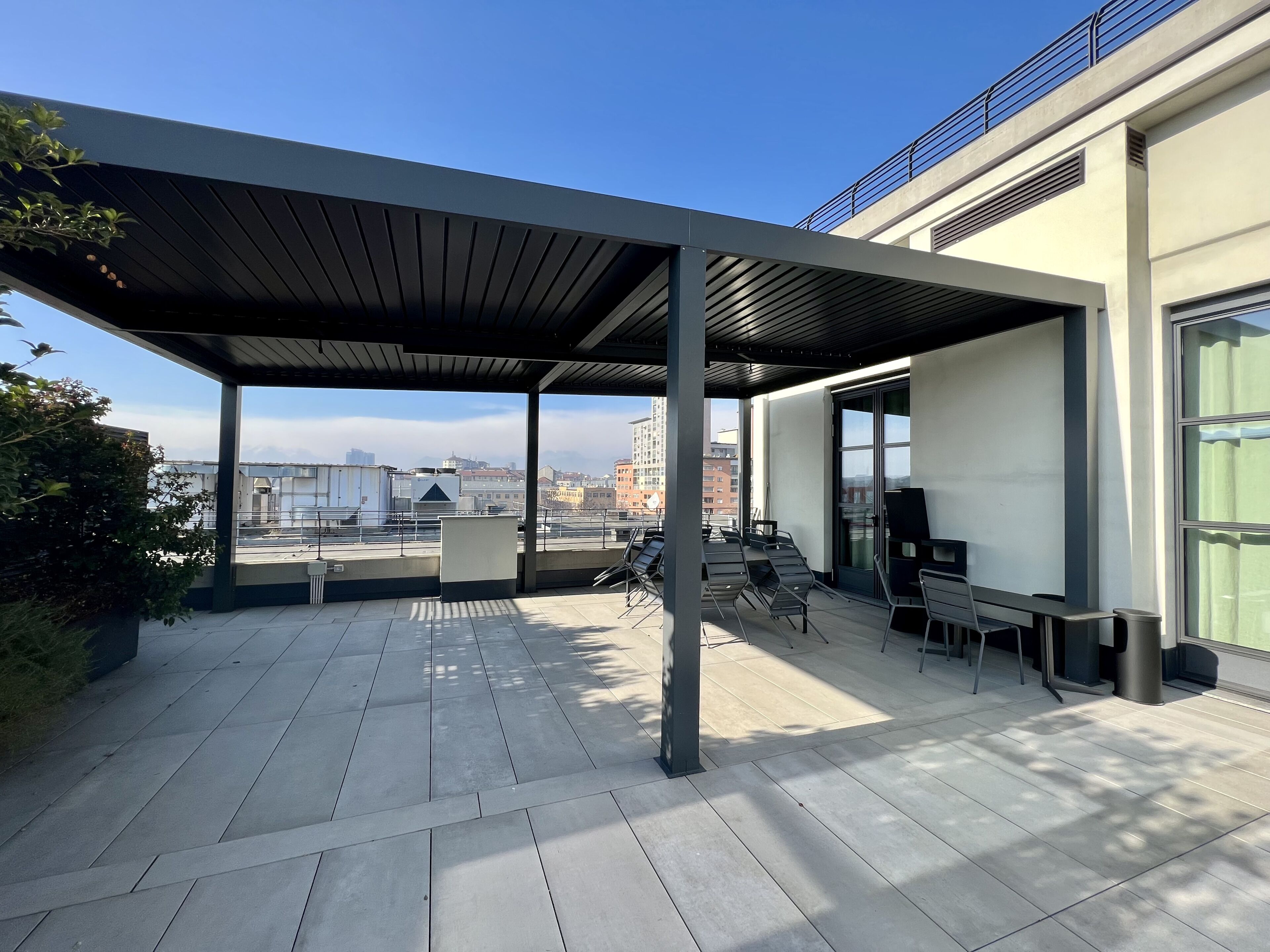 Una terraza en la azotea con zona de estar cubierta, vistas a la ciudad y un edificio moderno con grandes ventanales.