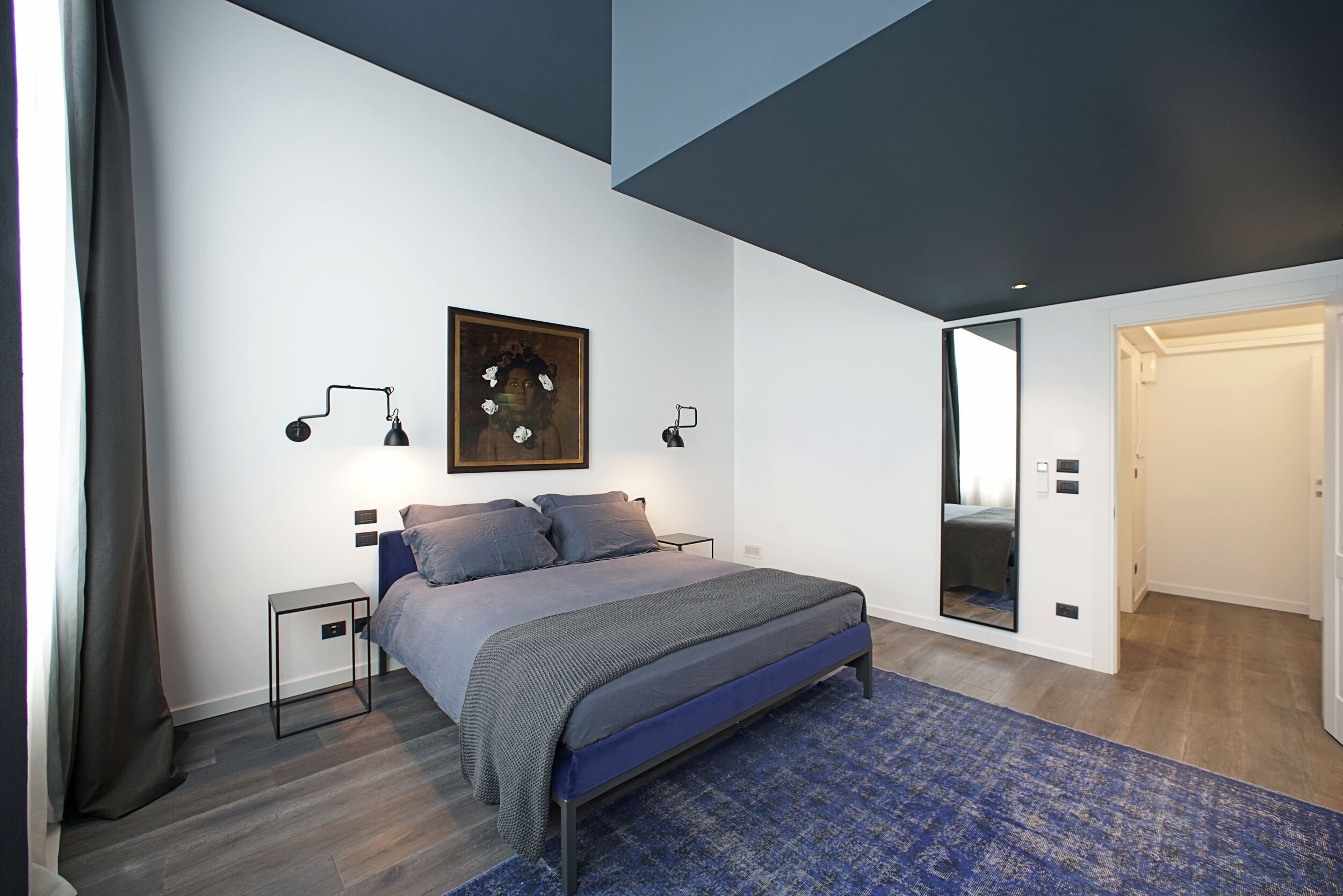 A modern bedroom with a bed, bedside lamps, a painting, and a mirror.