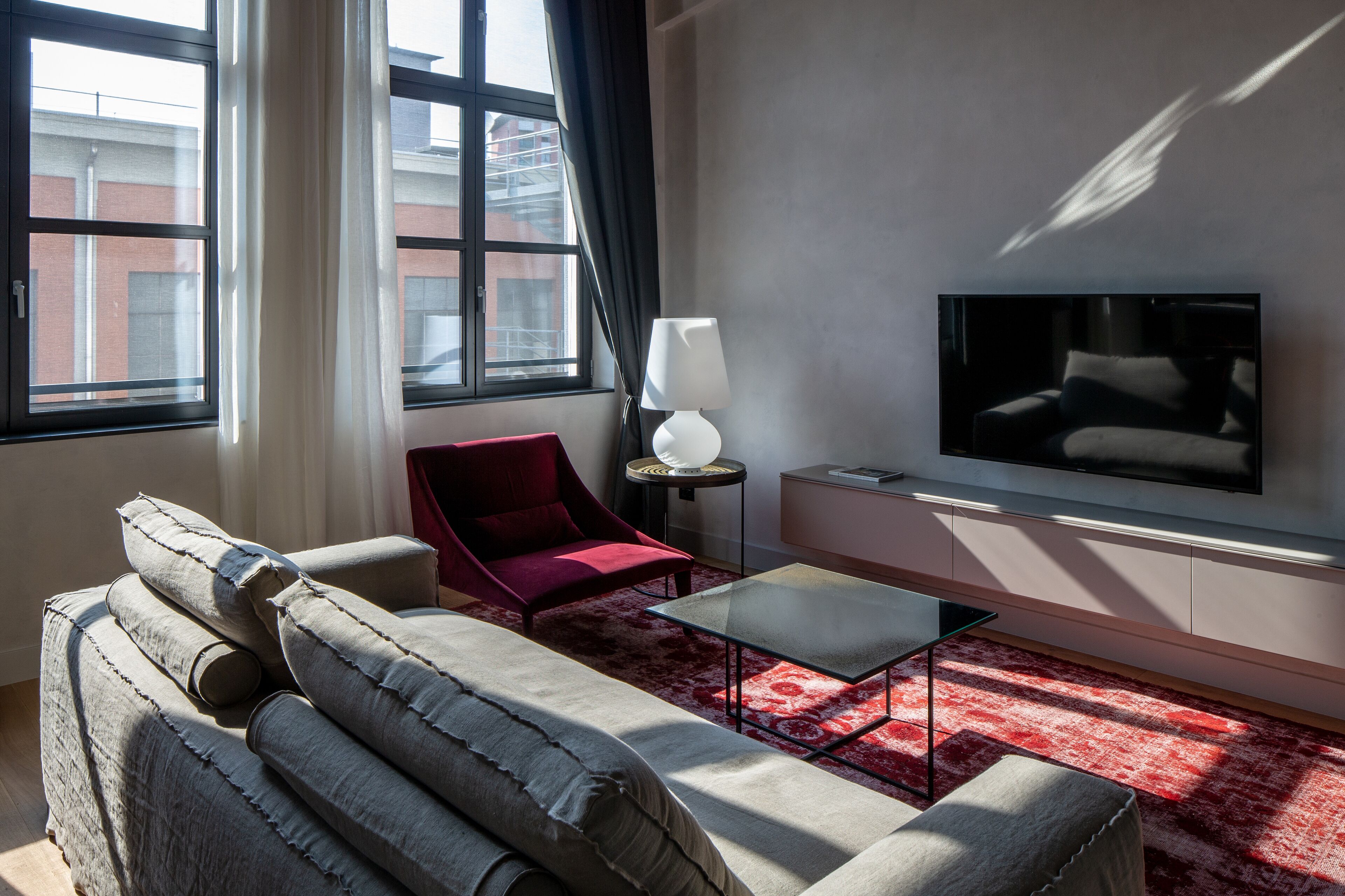 Uma sala de estar moderna com sofá, poltrona e mesa de centro. Uma TV de tela plana está fixada na parede, e um abajur sobre uma mesinha lateral.