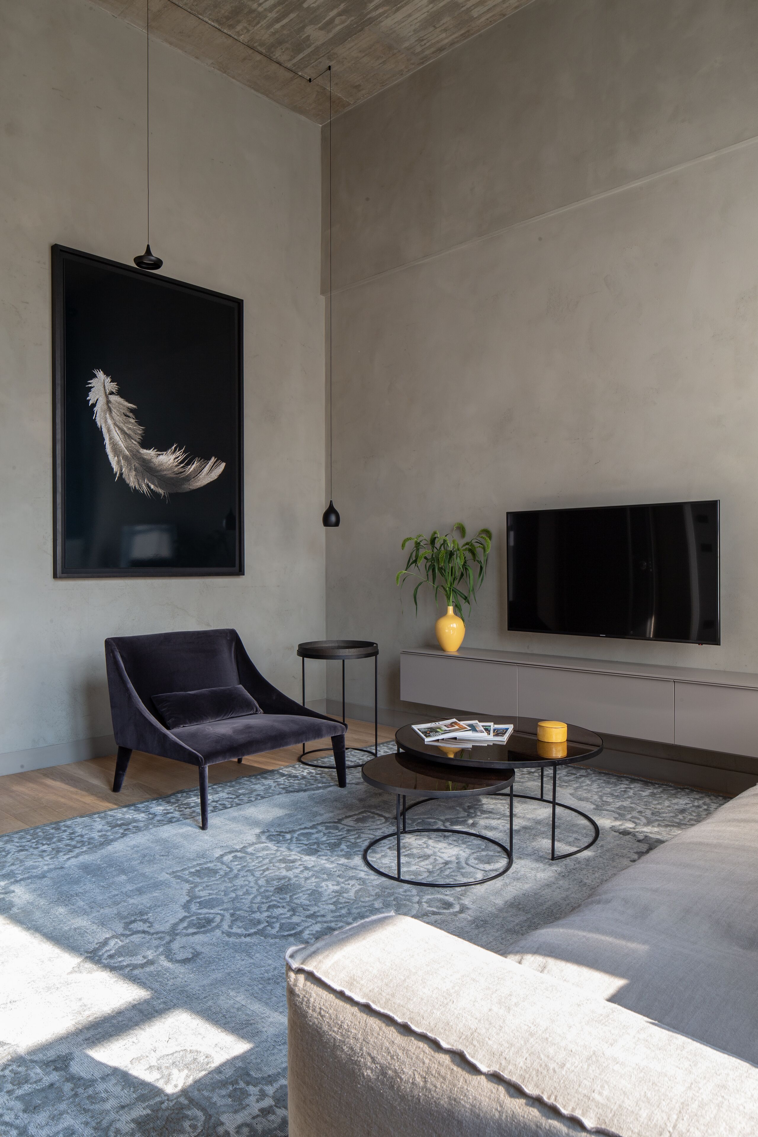 A modern living room with a large black feather artwork, a dark armchair, a round coffee table with magazines, a yellow vase with a plant, and a flat-screen TV on the wall.