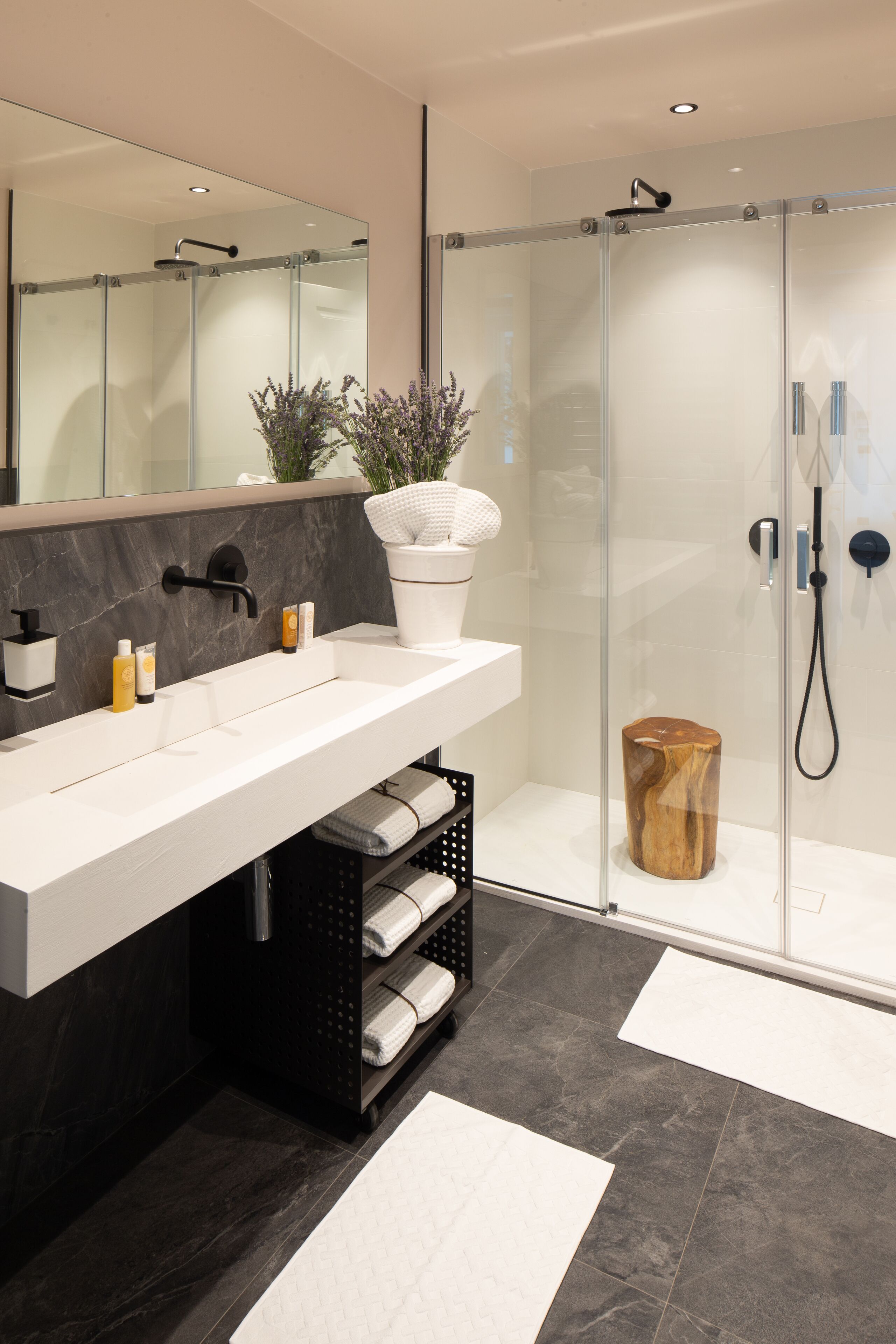 Une salle de bain moderne avec un grand miroir, un lavabo blanc, des carreaux foncés, une douche en verre et un tabouret en bois.